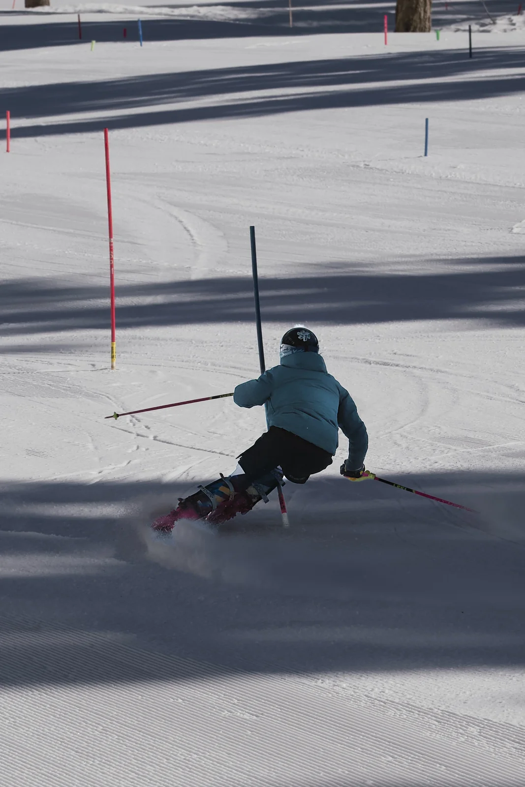Skier in a teal jacket carving a sharp turn through slalom gates, sending up a spray of snow