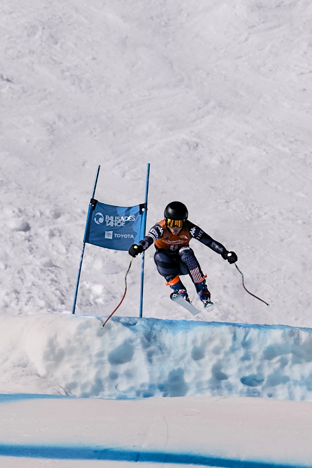 US team skier in an orange bib launching off a ridge past a Palisades Tahoe giant slalom gate