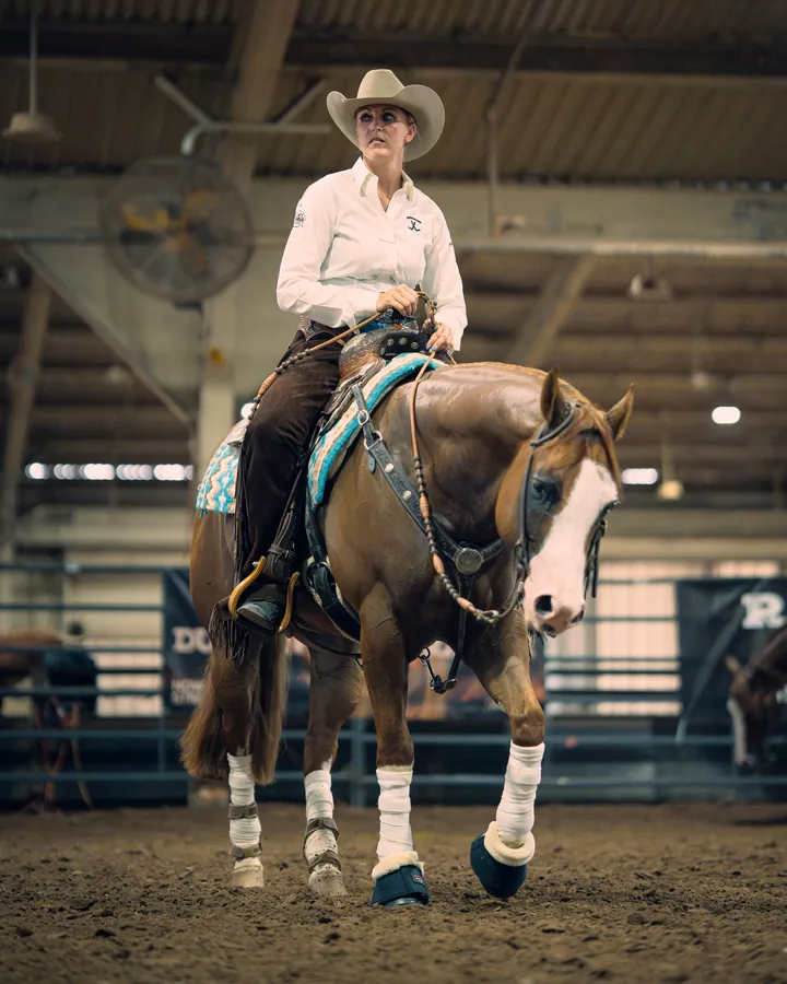 Female reining rider in a white shirt and cowboy hat aboard a chestnut horse with white leg wraps walking through an arena