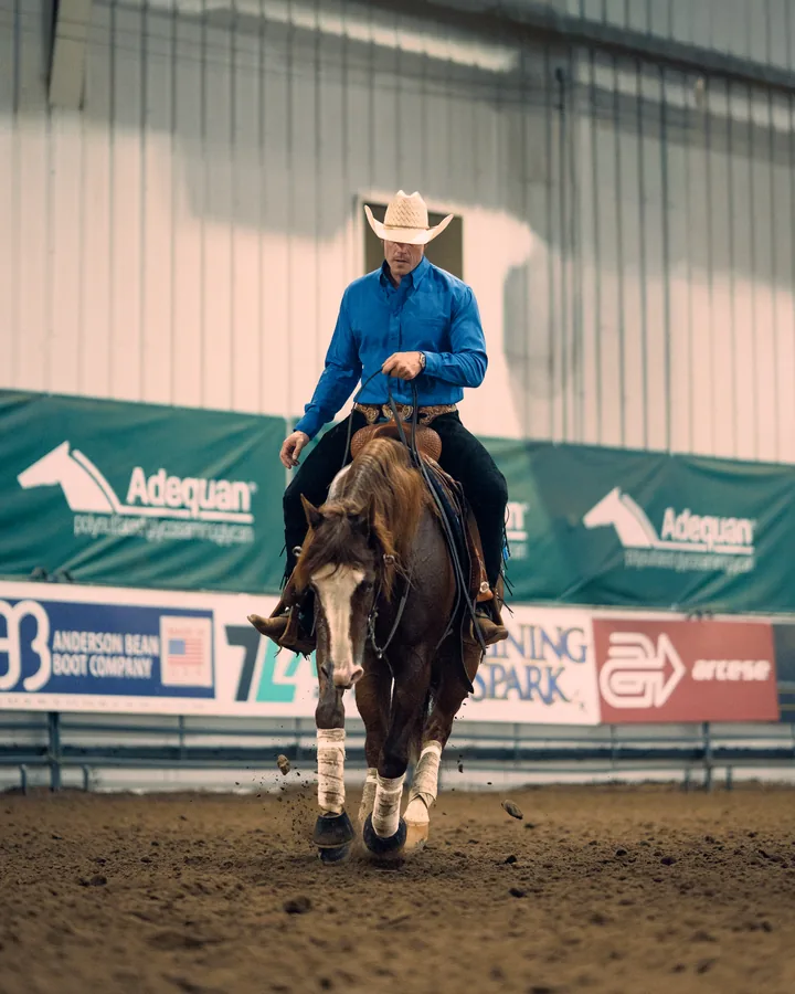 Cowboy in a blue shirt and straw hat riding a chestnut horse at a lope past sponsor banners in an indoor arena