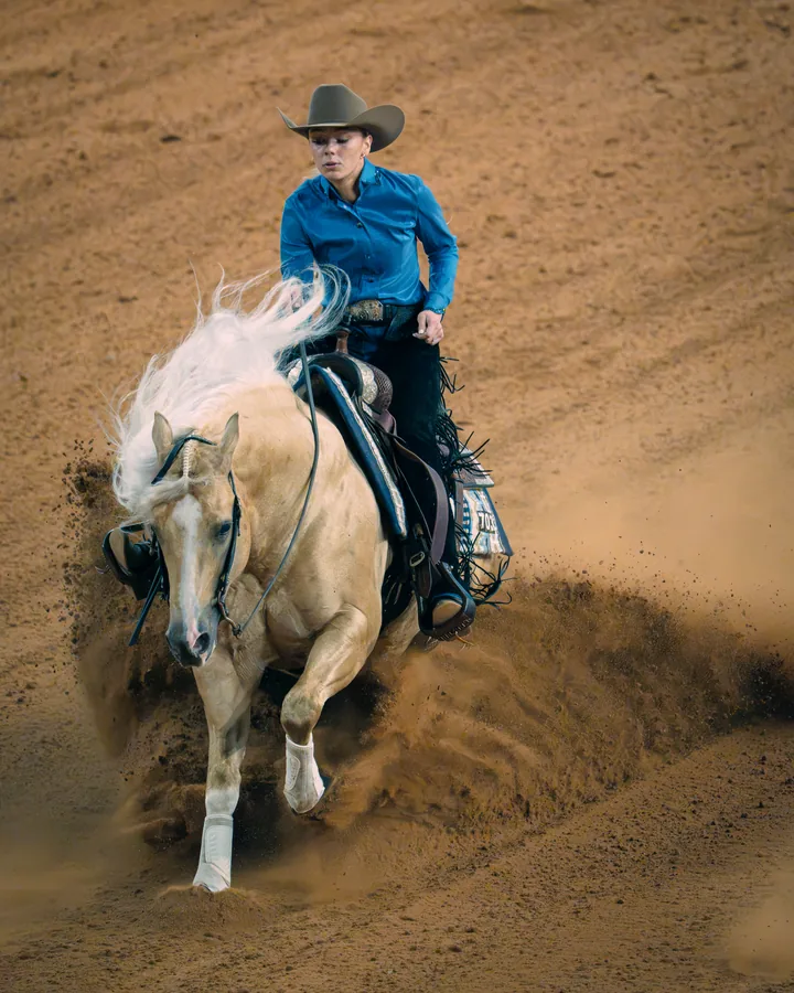 Female reiner in a teal shirt spinning a palomino horse in a dramatic sliding stop with dirt spraying in the arena