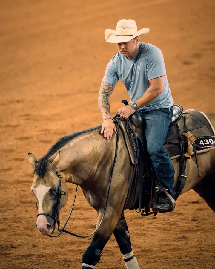 Tattooed cowboy in a light blue t-shirt and straw hat leaning forward to pat a buckskin horse in the arena