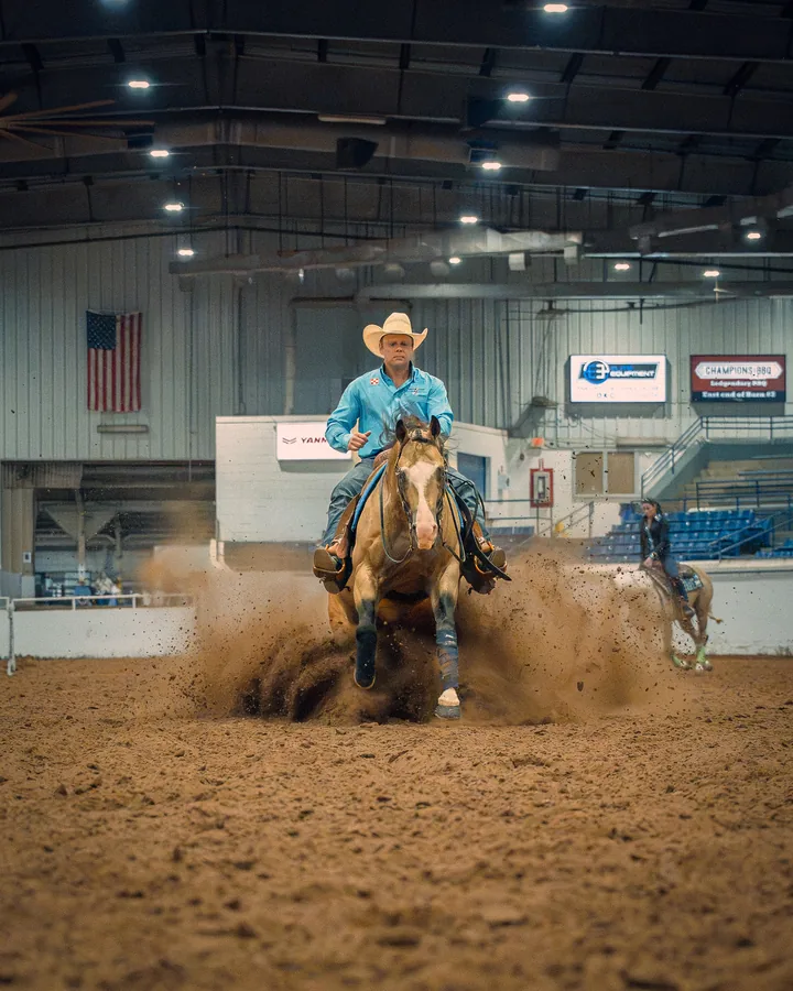 Cowboy in a turquoise shirt riding a paint horse sliding to a stop in a cloud of arena dust with an American flag in the background