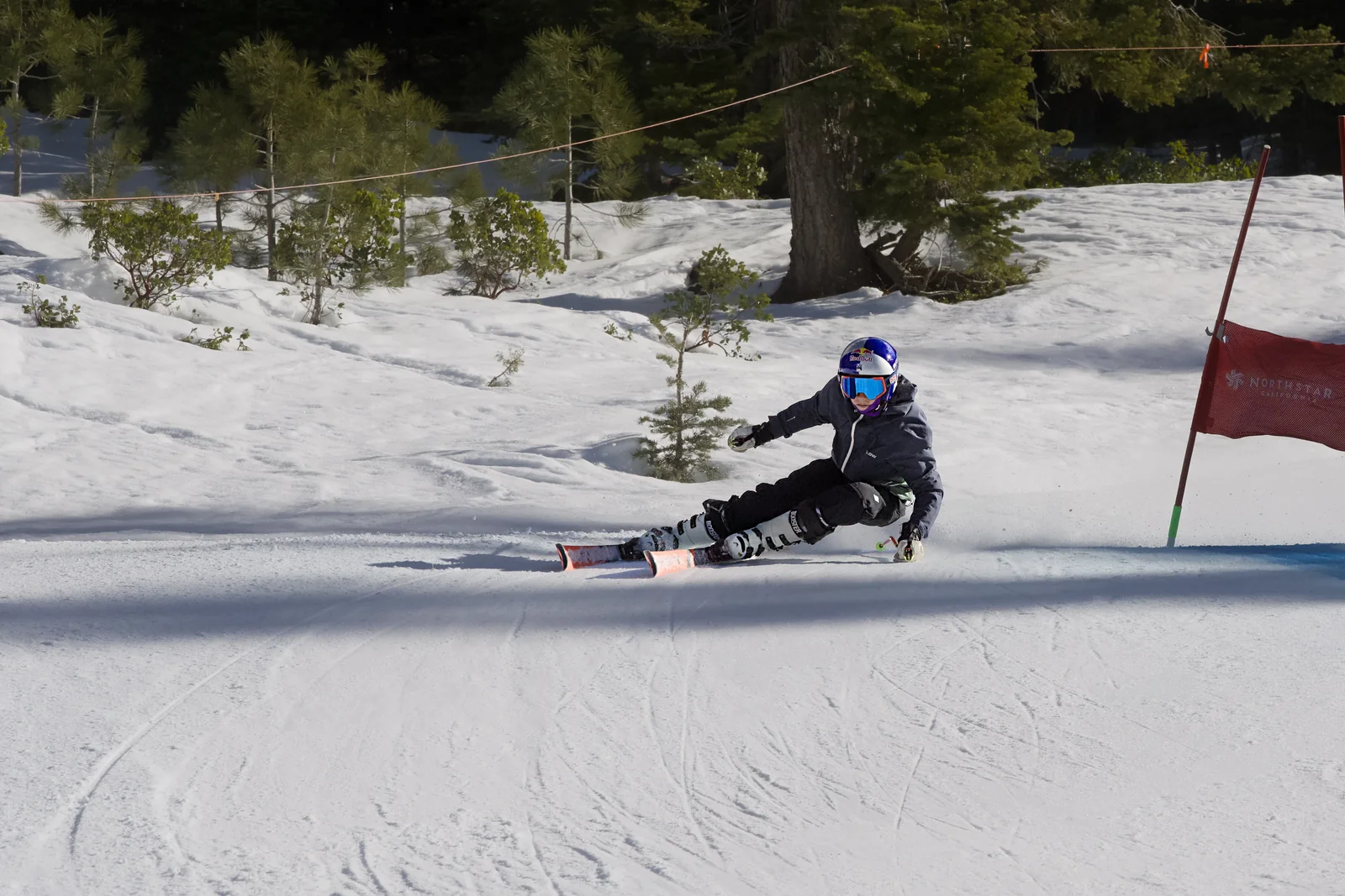 Skier in a dark suit making a deep low carve past a red gate on a groomed slope