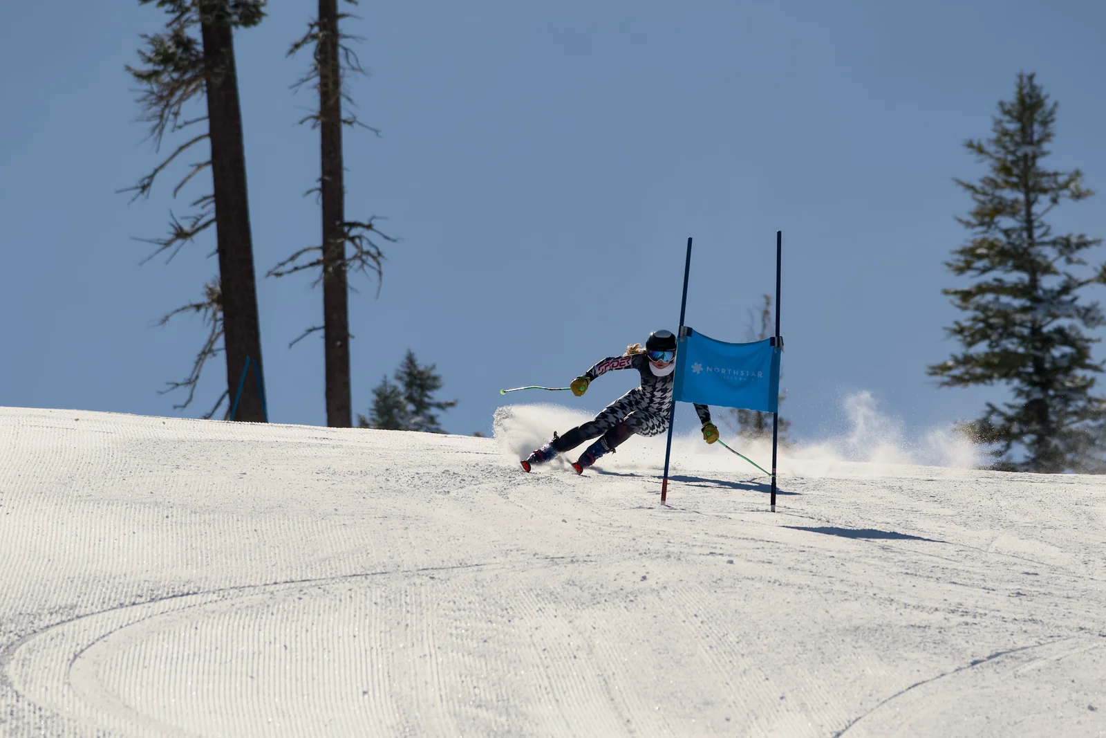 Skier in a houndstooth race suit ripping through a Northstar giant slalom gate in a cloud of snow