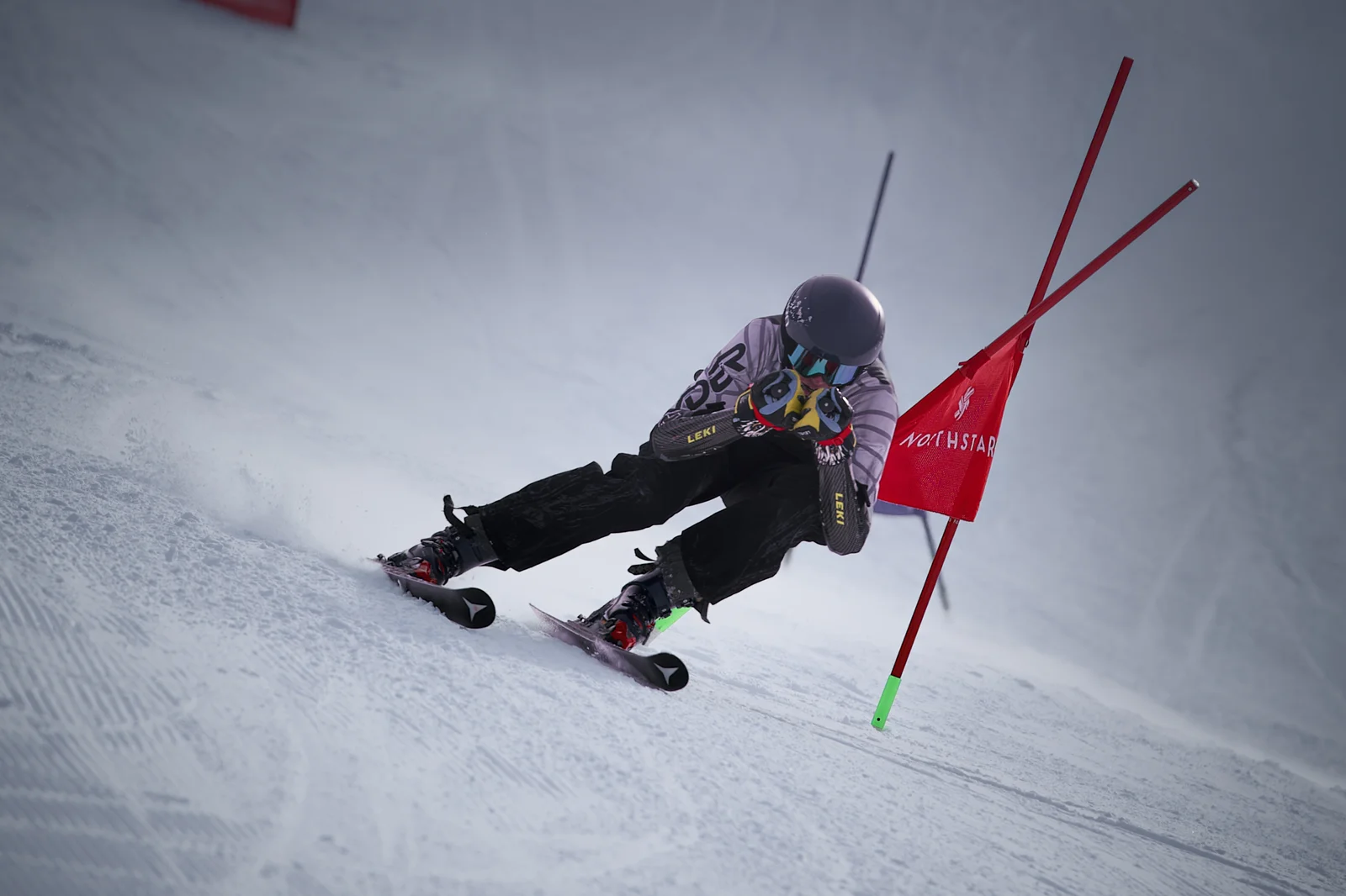 Skier in a dark grey suit driving low through a red Northstar giant slalom gate with snow spraying